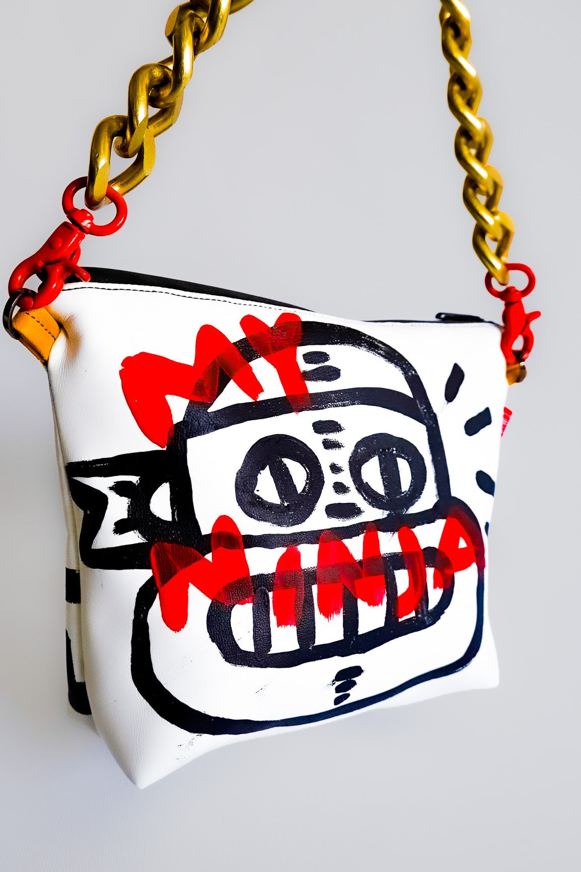 A white shoulder bag with a gold chain strap, featuring a black and red graffiti-style robot face graphic on the front.