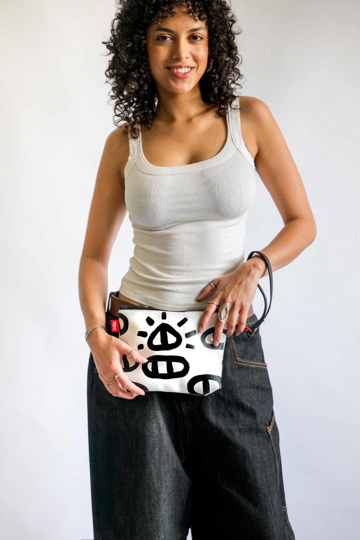 A woman with curly dark hair wearing a white tank top and dark jeans holds a white clutch bag decorated with black abstract symbols and a red button detail, set against a plain light gray background.