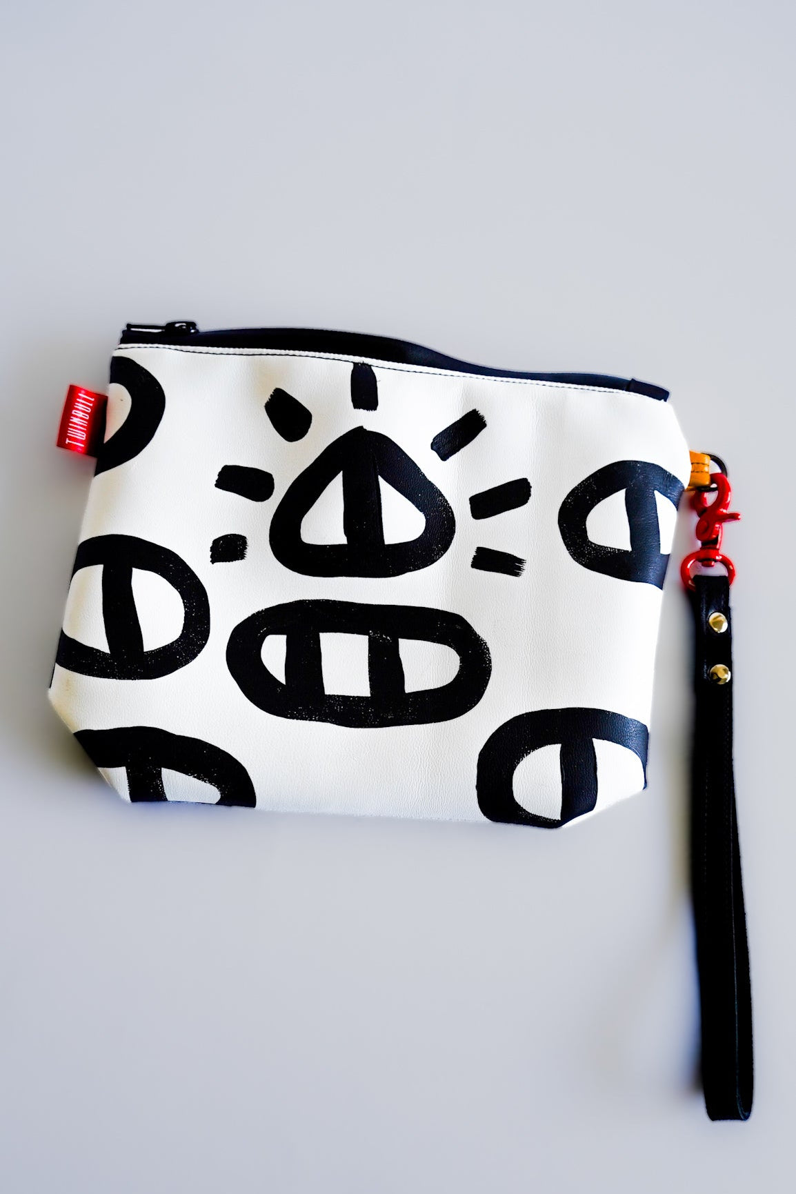 A white zippered pouch with a black hand-drawn abstract pattern, featuring a black wristlet strap with gold hardware and a red brand tag on a light gray background.