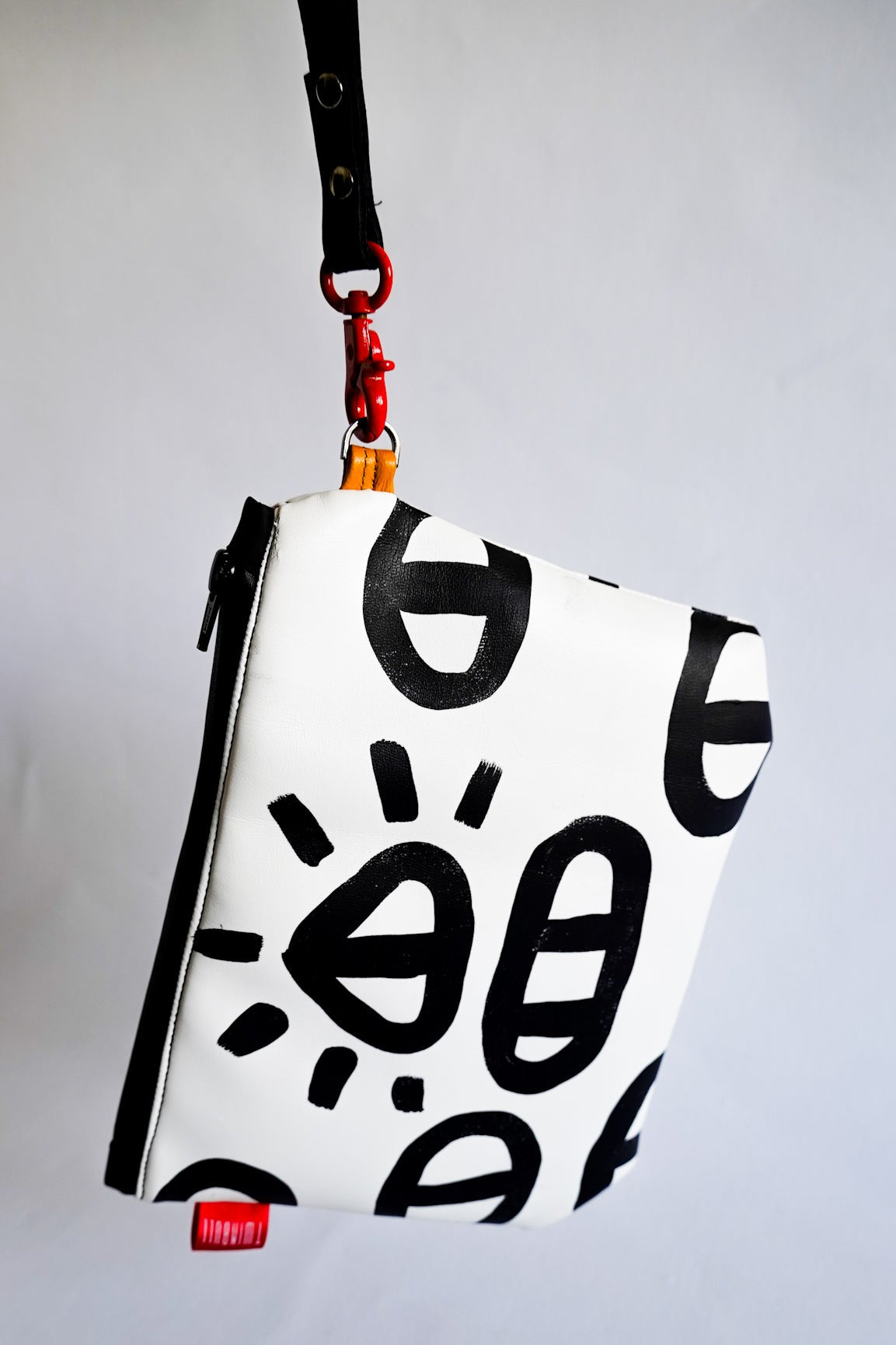 A white zippered pouch with a black Keith Haring style graphic pattern, featuring a black strap and red hardware, shown against a plain light gray background.