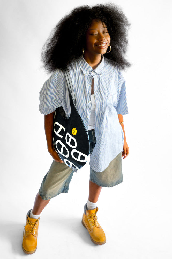 A woman with voluminous dark curly hair stands against a white studio background, wearing a light blue oversized button-up shirt, denim shorts, yellow boots, and holding a black tote bag with white graphic lettering.